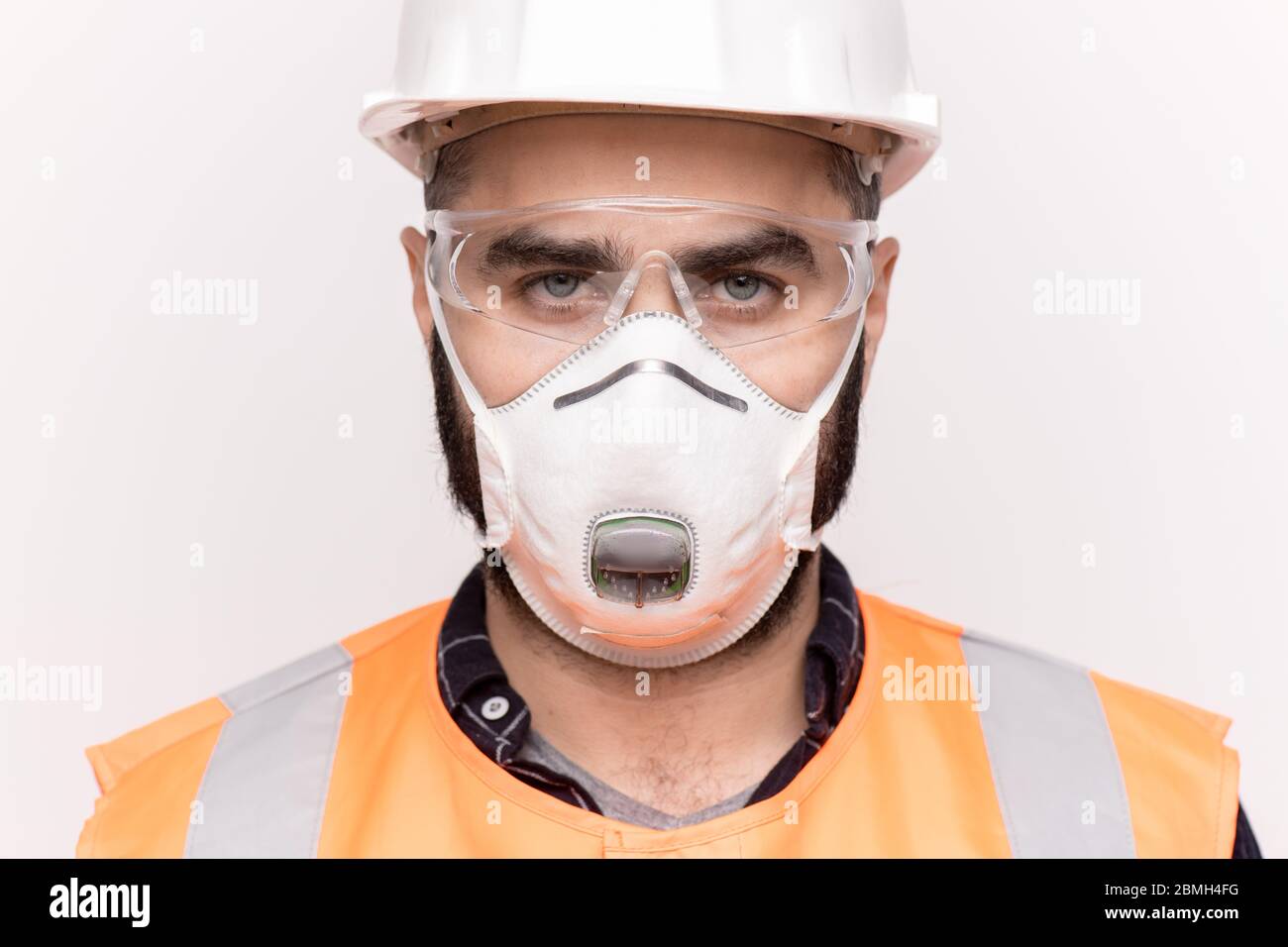 Un technicien professionnel sérieux en vêtements de travail orange, casque de protection, lunettes et masque vous regardant sur fond blanc Banque D'Images