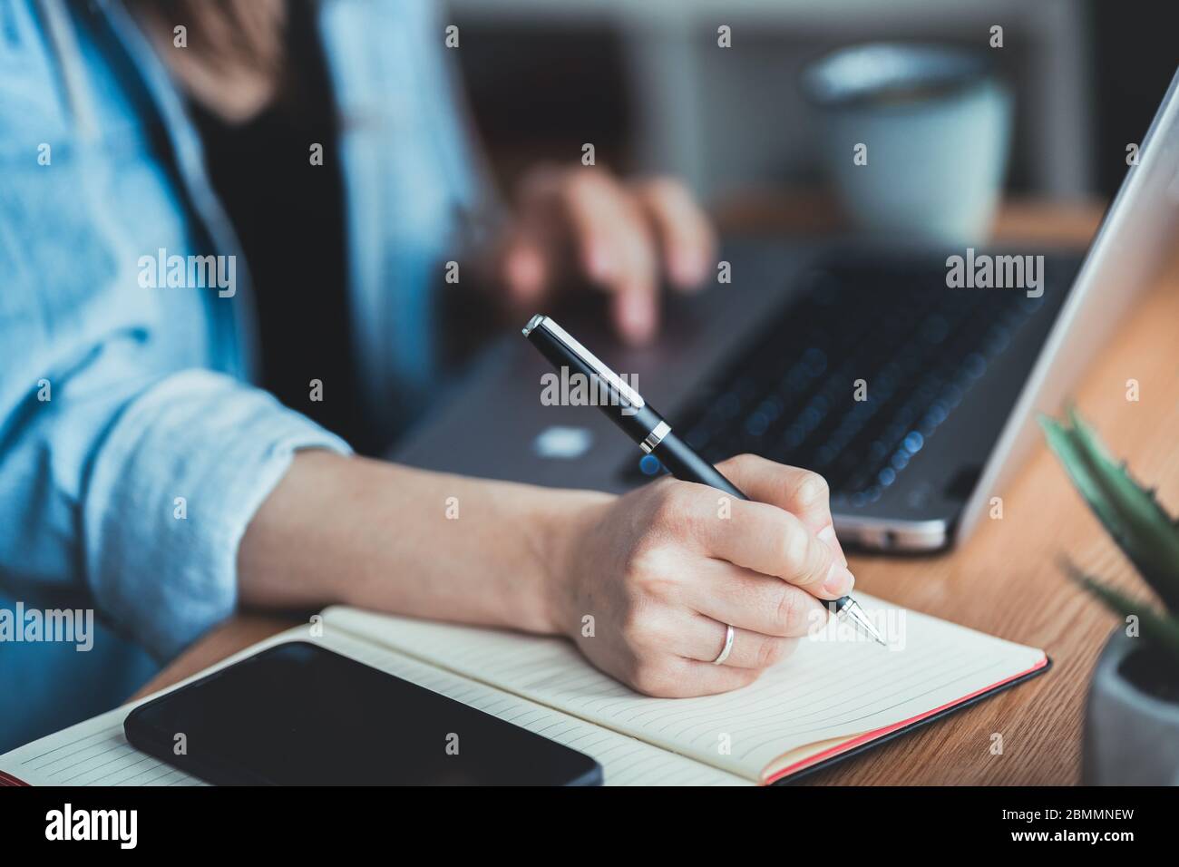 Jeune femme d'affaires assise au bureau, défilant sur un ordinateur portable et prenant des notes dans un ordinateur portable. Gros plan de l'écriture manuelle avec un stylo Banque D'Images