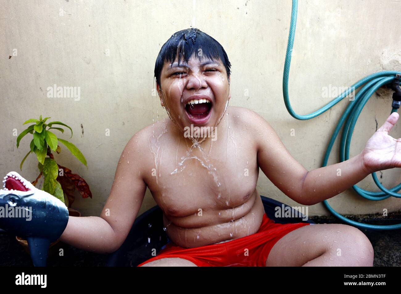Photo d'un jeune garçon asiatique qui se rafraîchi dans un bassin d'eau et un tuyau d'eau comme une douche de fortune pour battre la chaleur estivale. Banque D'Images