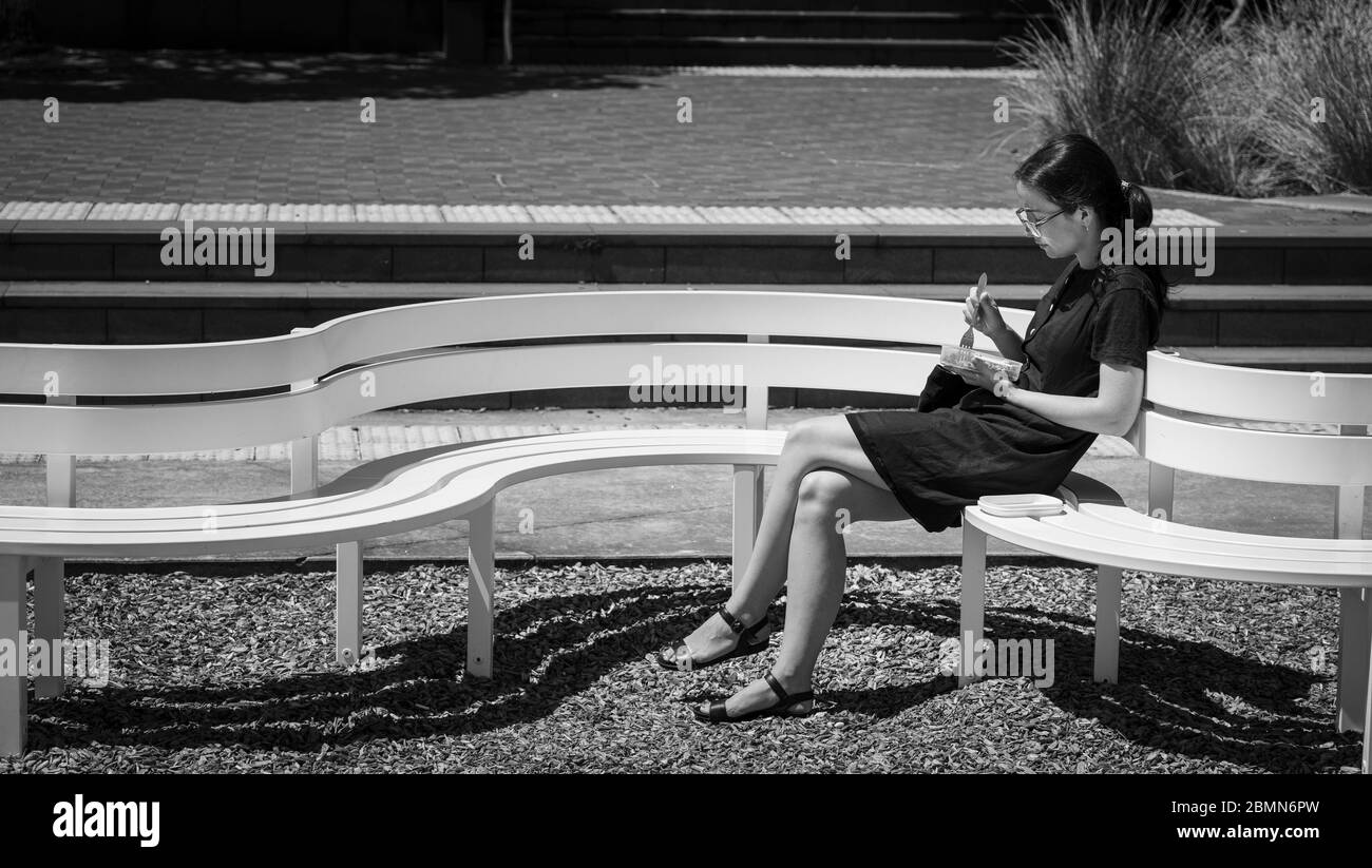 Une jeune femme asiatique se trouve au soleil sur un banc blanc incurvé pour déjeuner à Auckland, en Nouvelle-Zélande. Banque D'Images