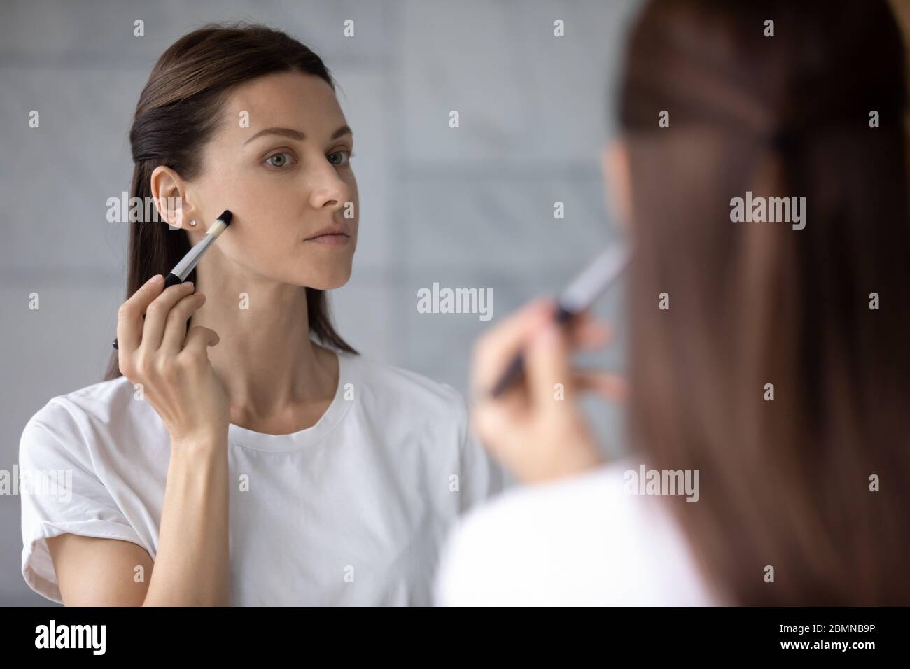 Femme reflétée dans le miroir tenant la brosse appliquant la crème de correcteur de fondation Banque D'Images