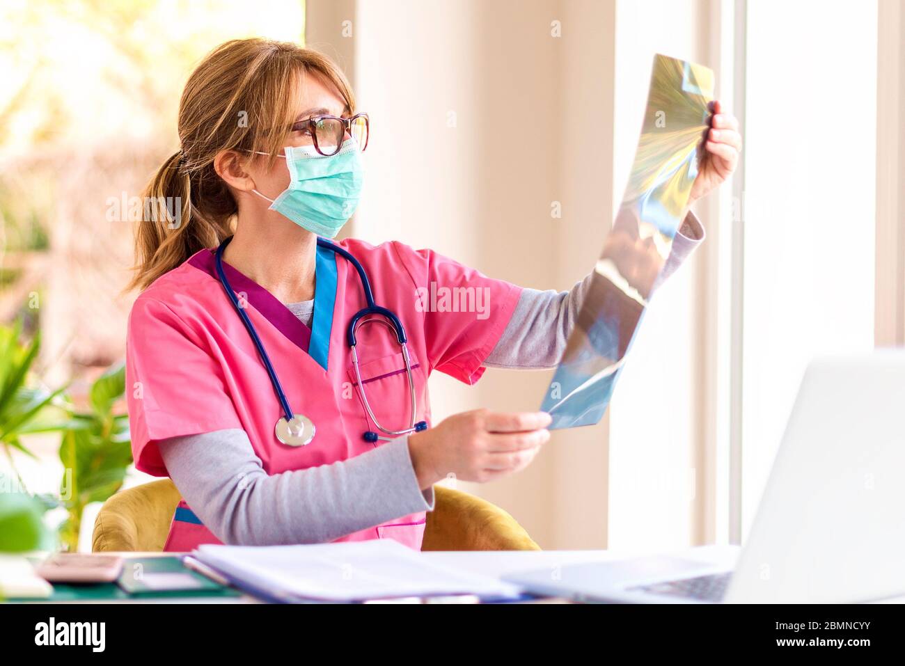 Prise de vue d'un médecin portant un masque facial assis dans la salle du médecin et vérifiant l'image radiographique. Banque D'Images