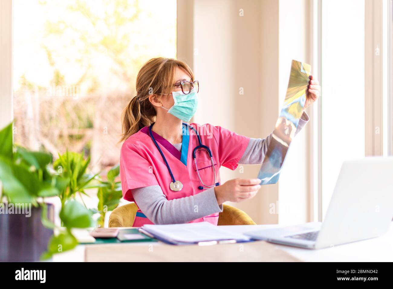 Prise de vue d'un médecin portant un masque facial assis dans la salle du médecin et vérifiant l'image radiographique. Banque D'Images