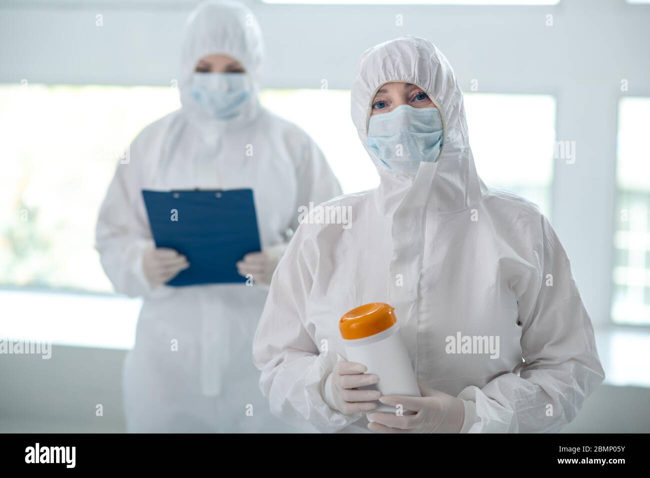 Travailleur médical en vêtements de protection tenant un flacon de lingettes désinfectantes, sa collègue se tenant derrière avec un dossier de clips Banque D'Images