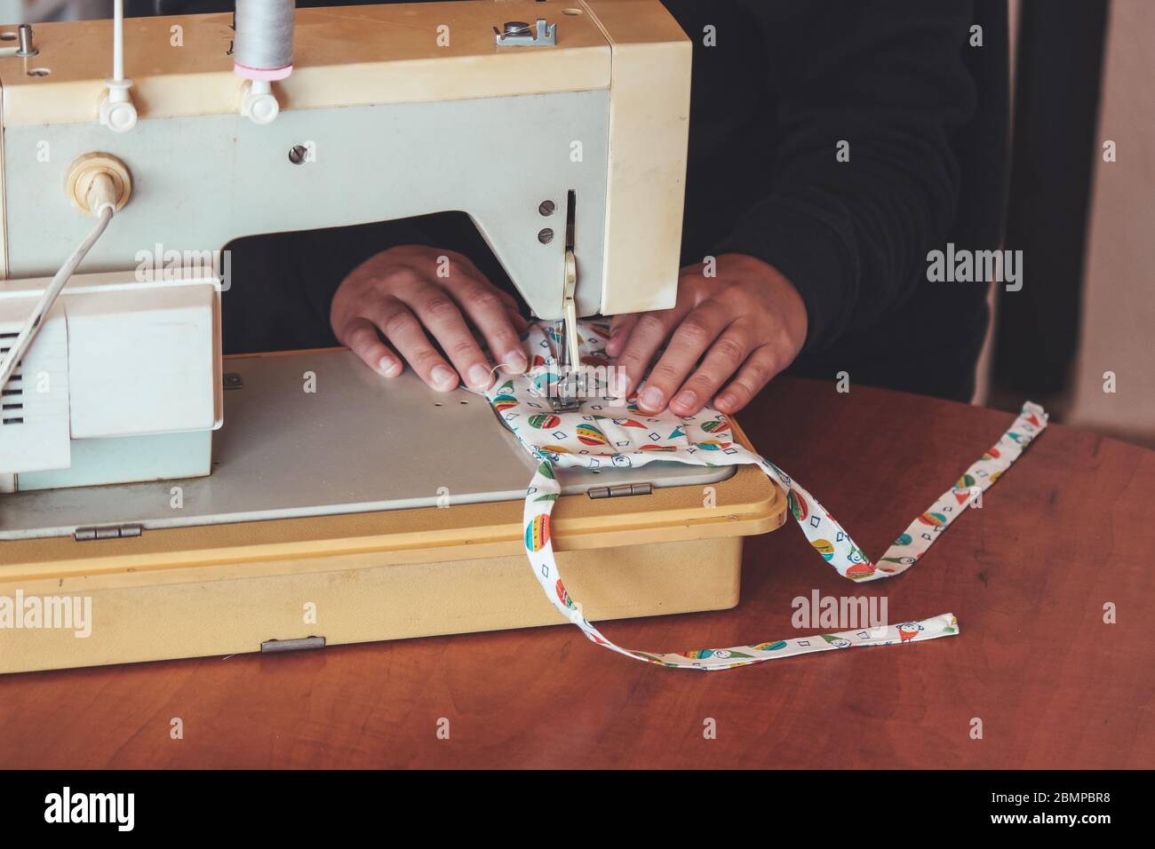 Les mains des femmes utilisant la machine à coudre pour coudre un masque médical coloré du visage pendant la pandémie de coronavirus. Masque de protection fait maison contre le virus. Masques à coudre, COVID-19. Mise au point sélective. Banque D'Images