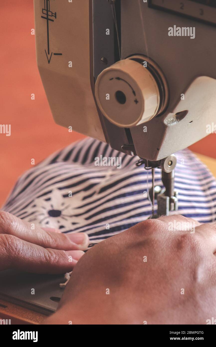 Les mains des femmes utilisant la machine à coudre pour coudre un masque médical coloré du visage pendant la pandémie de coronavirus. Masque de protection fait maison contre le virus. Masques à coudre, COVID-19. Mise au point sélective. Banque D'Images