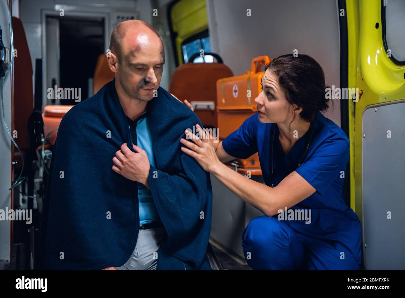 Une jeune femme dans un uniforme médical bleu essayant de réconforter un homme blessé, assis sur le bord d'une voiture d'ambulance dans une couverture Banque D'Images