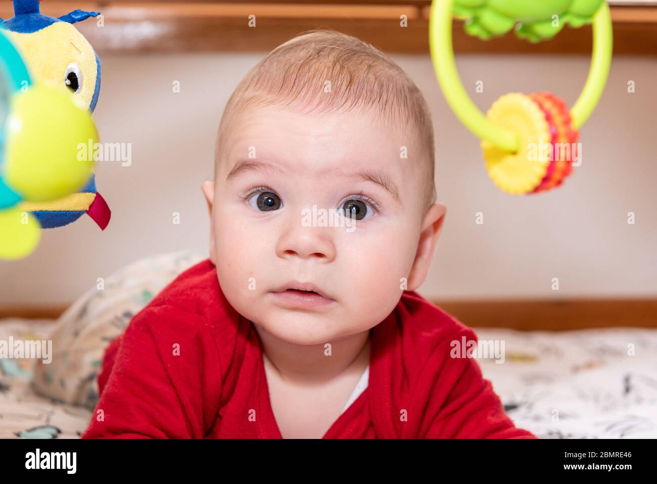 Mignon petit garçon pendant le ventre regarder l'appareil photo, enfant de 6 mois avec la curiosité expression sur son visage entouré de jouets colorés. Banque D'Images