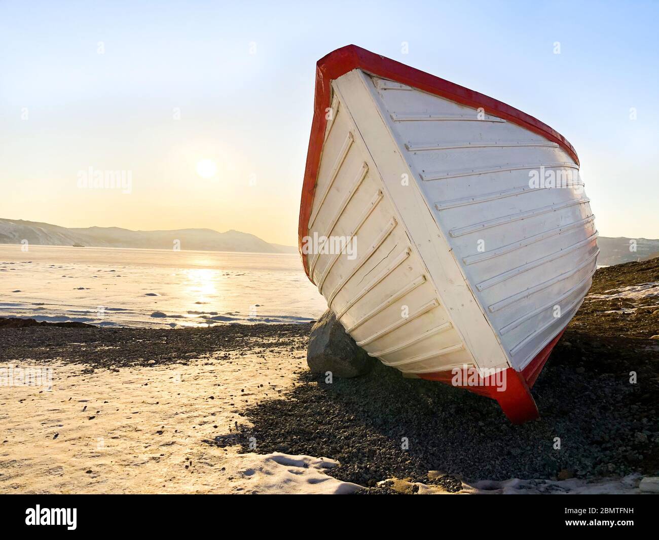 Photo d'un bateau en bois sur le bord de mer de Magadan, Russie. Bateau blanc à rayures rouges. Coucher de soleil sur l'arrière-plan. Ciel bleu clair et glace sur la mer en hiver Banque D'Images