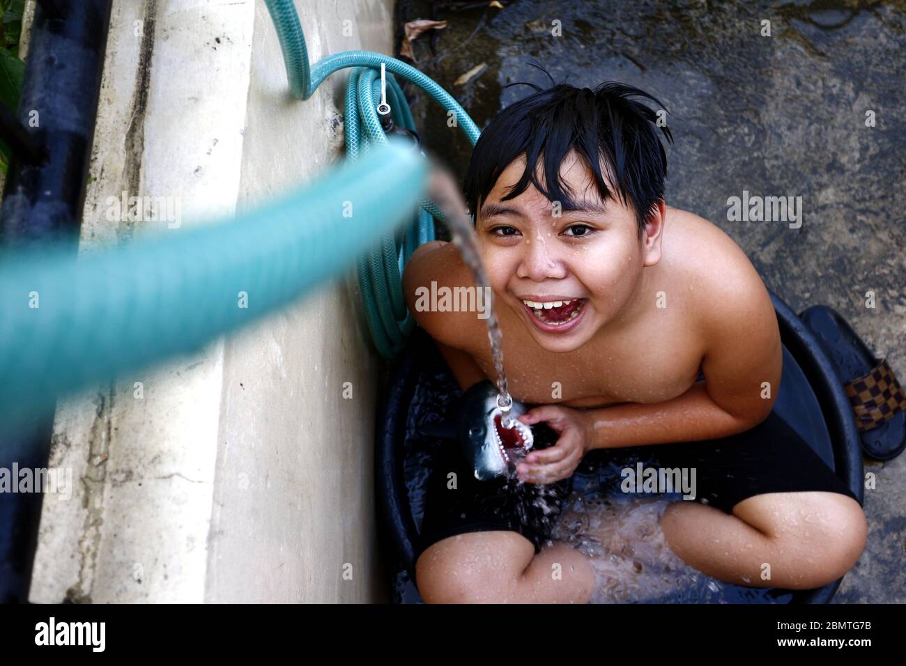Photo d'un jeune garçon asiatique qui se rafraîchi dans un bassin d'eau avec un tuyau d'eau comme une douche de fortune pour battre la chaleur estivale. Banque D'Images