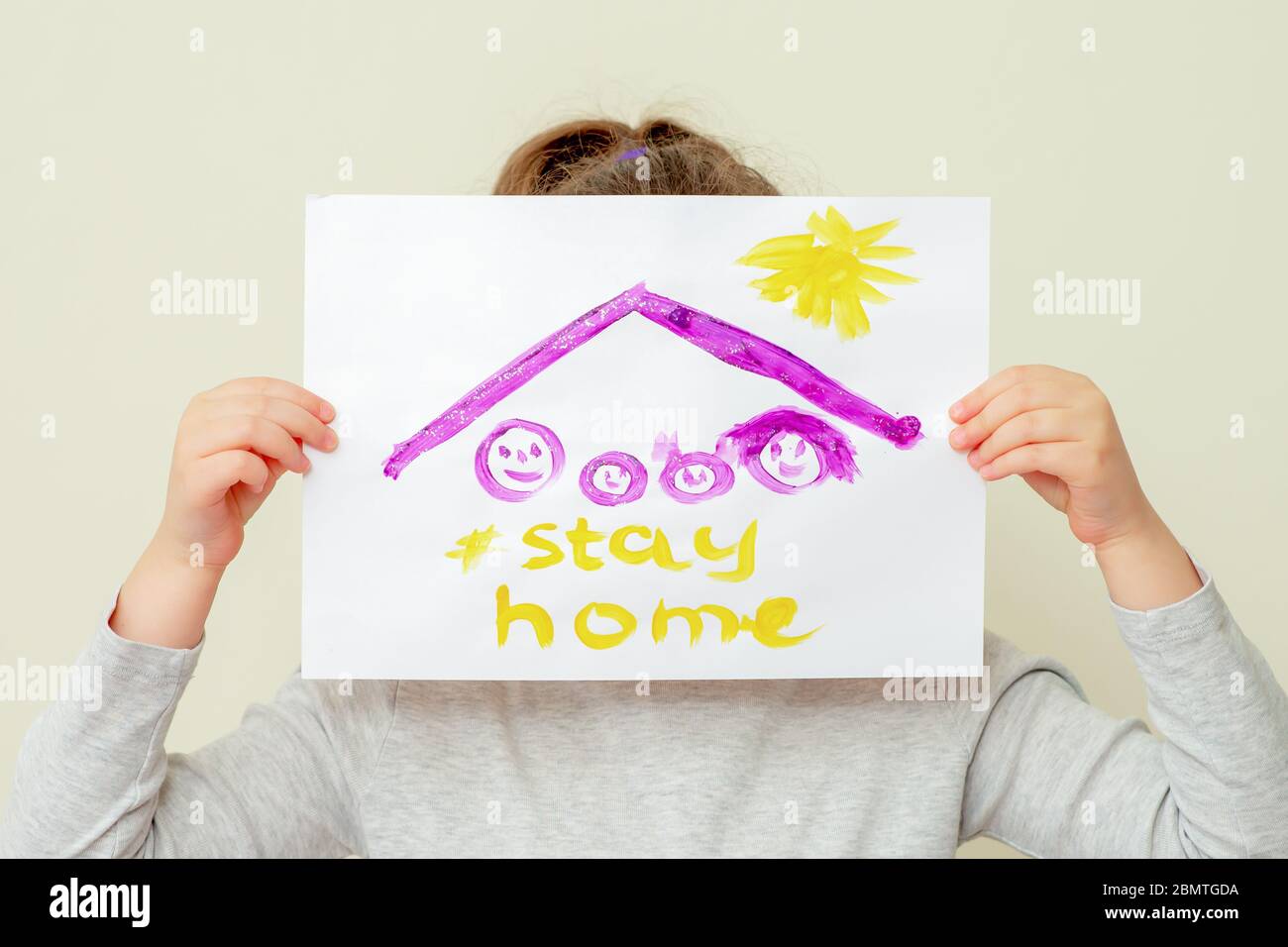 Mains de l'enfant tenant une photo de la silhouette de famille sous le toit et les mots rester à la maison couvrant son visage sur fond jaune. Enfants en quarantaine. Banque D'Images