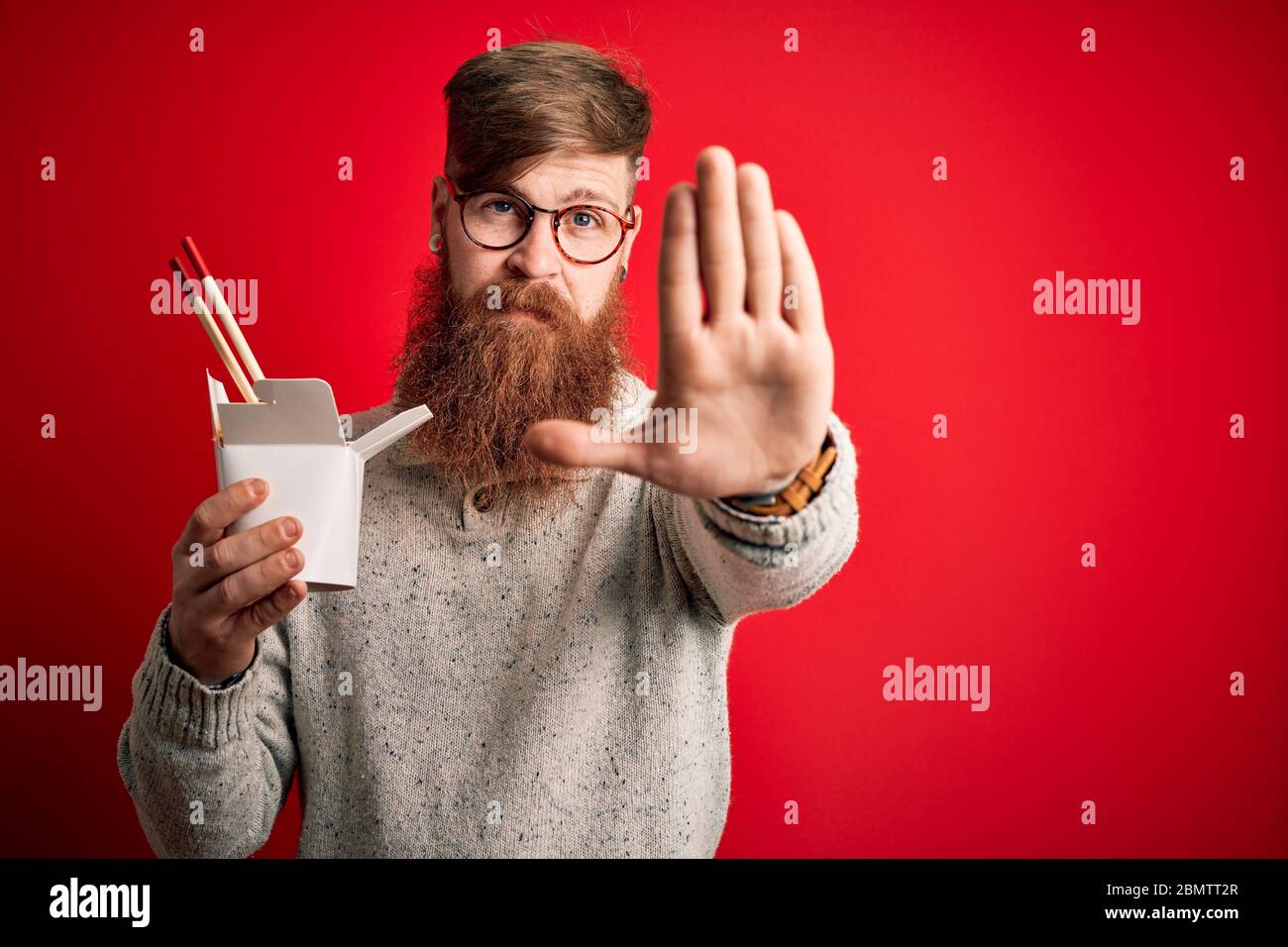 REDHEAD Irlandais homme avec la barbe boîte de ramen manger de la nourriture asiatique en utilisant des baguettes avec main ouverte faire signe d'arrêt avec sérieux et confiant express Banque D'Images