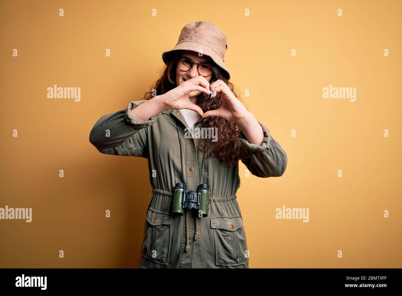 Jeune belle femme touristique en vacances portant un chapeau d'explorateur  et des jumelles souriant dans l'amour faisant le symbole de forme de coeur  avec les mains. Concept romantique Photo Stock - Alamy
