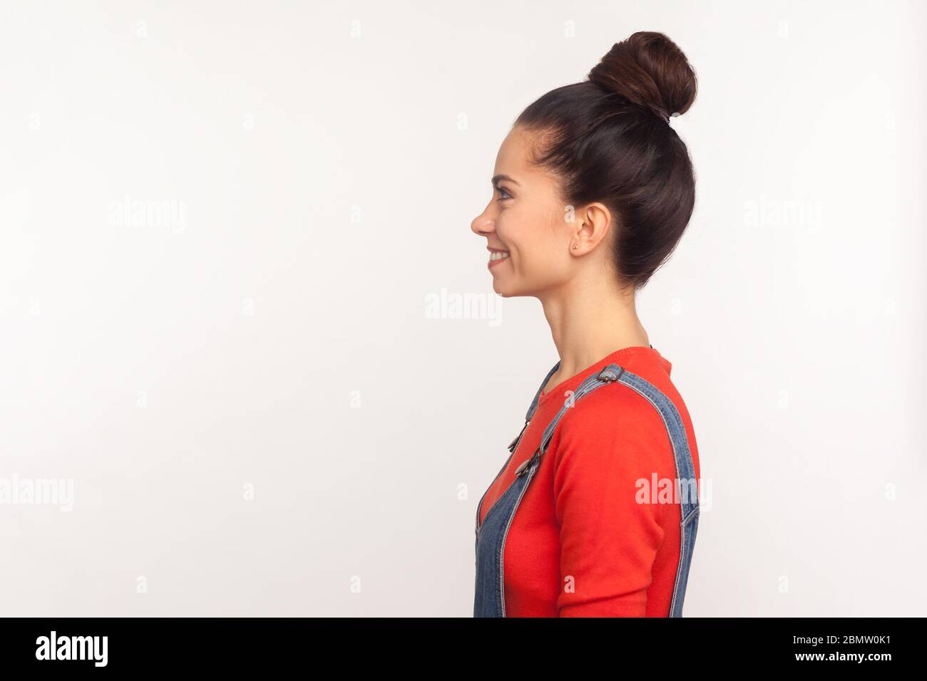 Profil de positive heureux stylisé jolie fille avec un pain de cheveux dans des combinaisons en denim regardant sur le côté de l'espace de copie avec le sourire en dents de souris, exprimant la joie et l'optimiste Banque D'Images