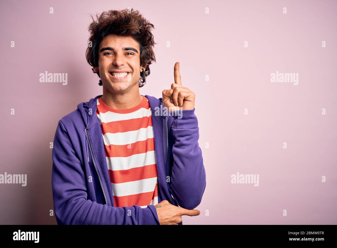 Jeune homme portant un t-shirt rayé et un sweat-shirt violet sur fond rose isolé avec un grand sourire sur le visage, pointant avec la main et le doigt vers le si Banque D'Images