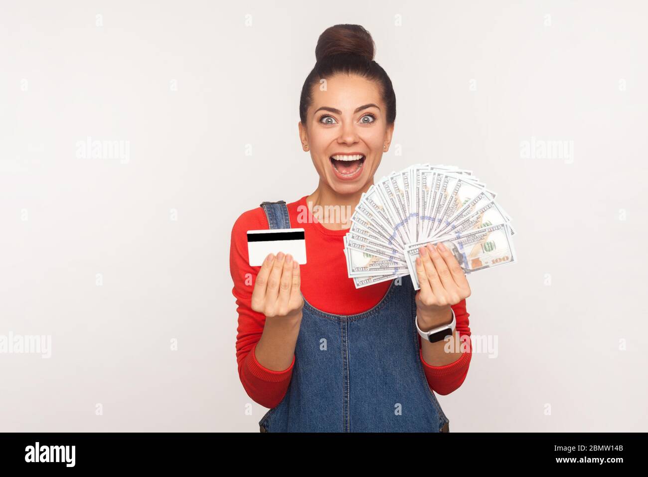 Wow, prêt bancaire, cashback! Portrait de la fille excitée avec cheveux chignon dans les combinaisons de denim tenant la carte de crédit et beaucoup d'argent, choqué par l'argent deposi Banque D'Images