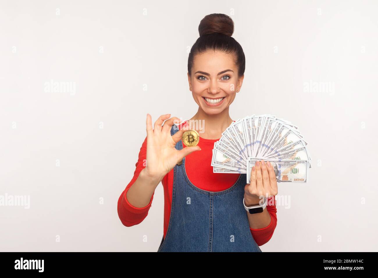 Investissements, argent numérique. Portrait d'une fille élégante et heureuse avec un pain en cheveux dans des combinaisons en denim contenant du bitcoin doré et des billets de banque en dollars, souriant à la came Banque D'Images