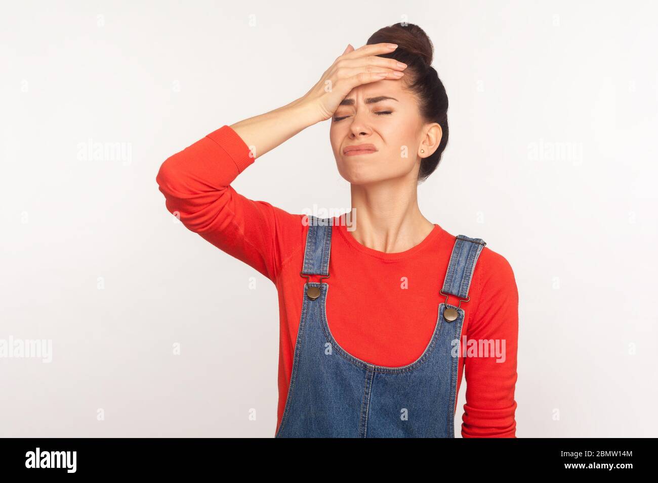 Dommage que j'ai oublié ! Portrait d'une fille inquiète avec un cheveu dans une combinaison en denim debout avec geste de visage, désespéré de mauvais souvenir, lui reprochant Banque D'Images