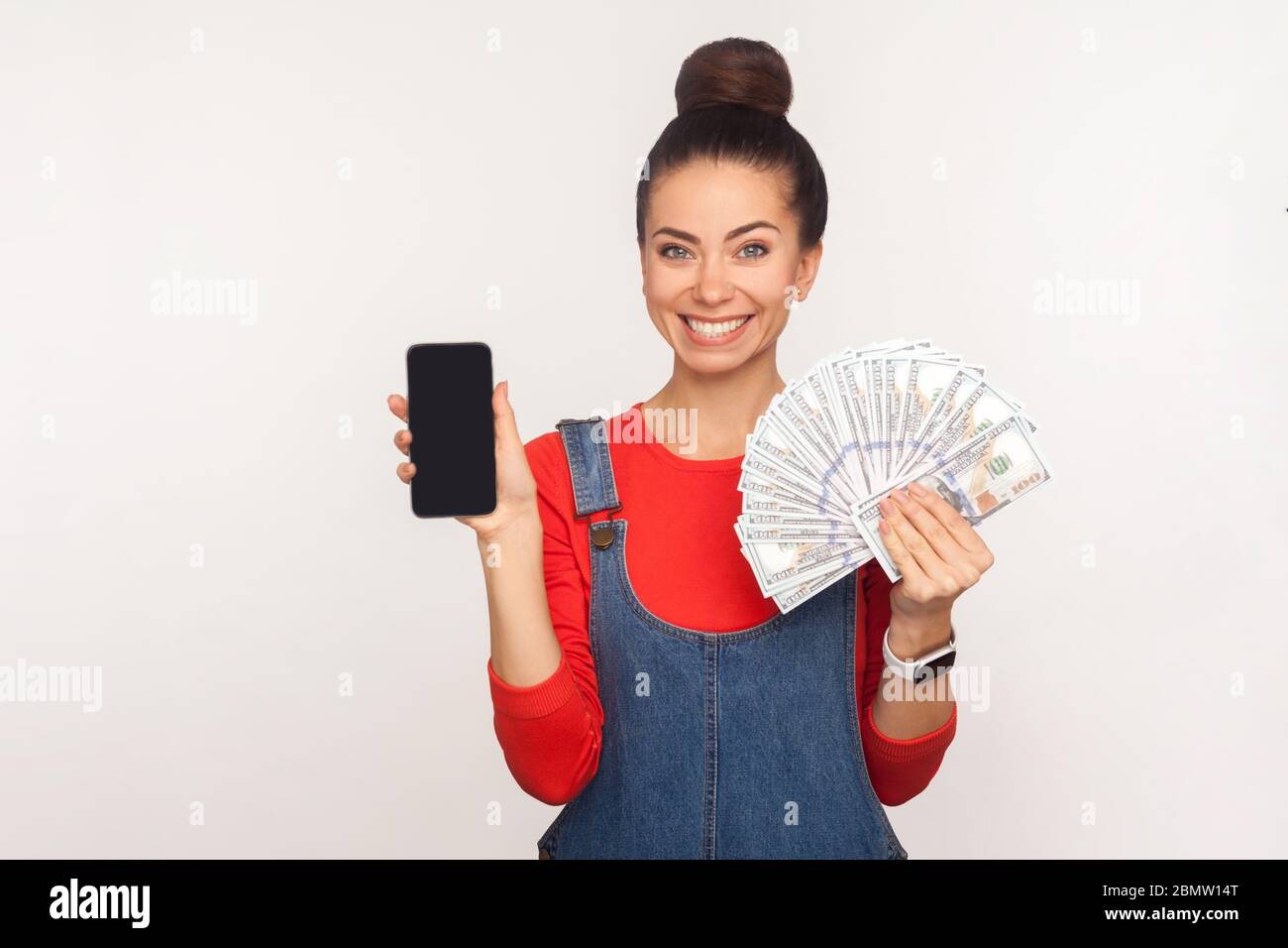 Paiement mobile, achats en ligne. Portrait de Happy élégant jolie fille avec cheveux chignon dans des combinaisons en denim tenant téléphone cellulaire et argent, billets de dollars. ind Banque D'Images