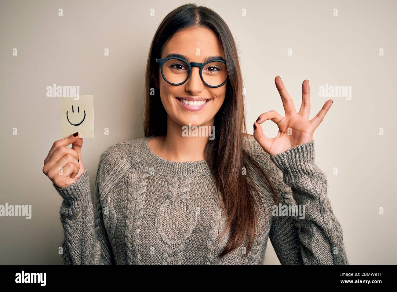 Jeune belle femme brune portant des lunettes tenant le papier avec sourire  emoji faisant ok signe avec les doigts, excellent symbole Photo Stock -  Alamy