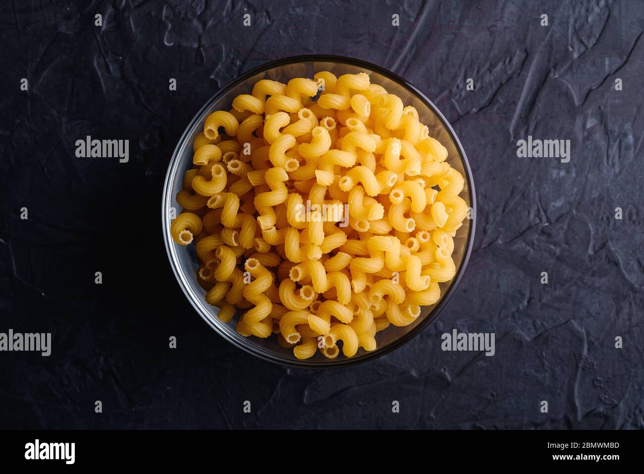 Bol en verre avec pâtes cavataappi au blé doré non cuites sur fond noir foncé texturé, vue du dessus Banque D'Images