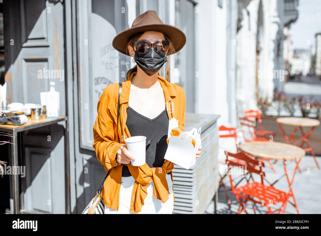 Portrait d'une jeune femme dans un masque de protection du visage debout avec une nourriture à emporter près de l'entrée du café à l'extérieur. Concept de nouvelles règles sociales pour les entreprises après une pandémie de coronavirus Banque D'Images