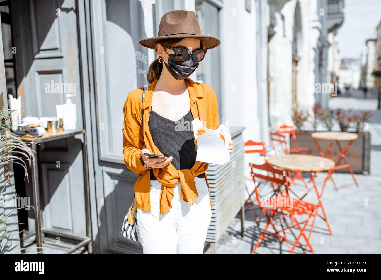 Jeune femme dans un masque de protection du visage debout avec une nourriture à emporter près de l'entrée du café à l'extérieur. Concept de nouvelles règles sociales pour les entreprises après une pandémie de coronavirus Banque D'Images