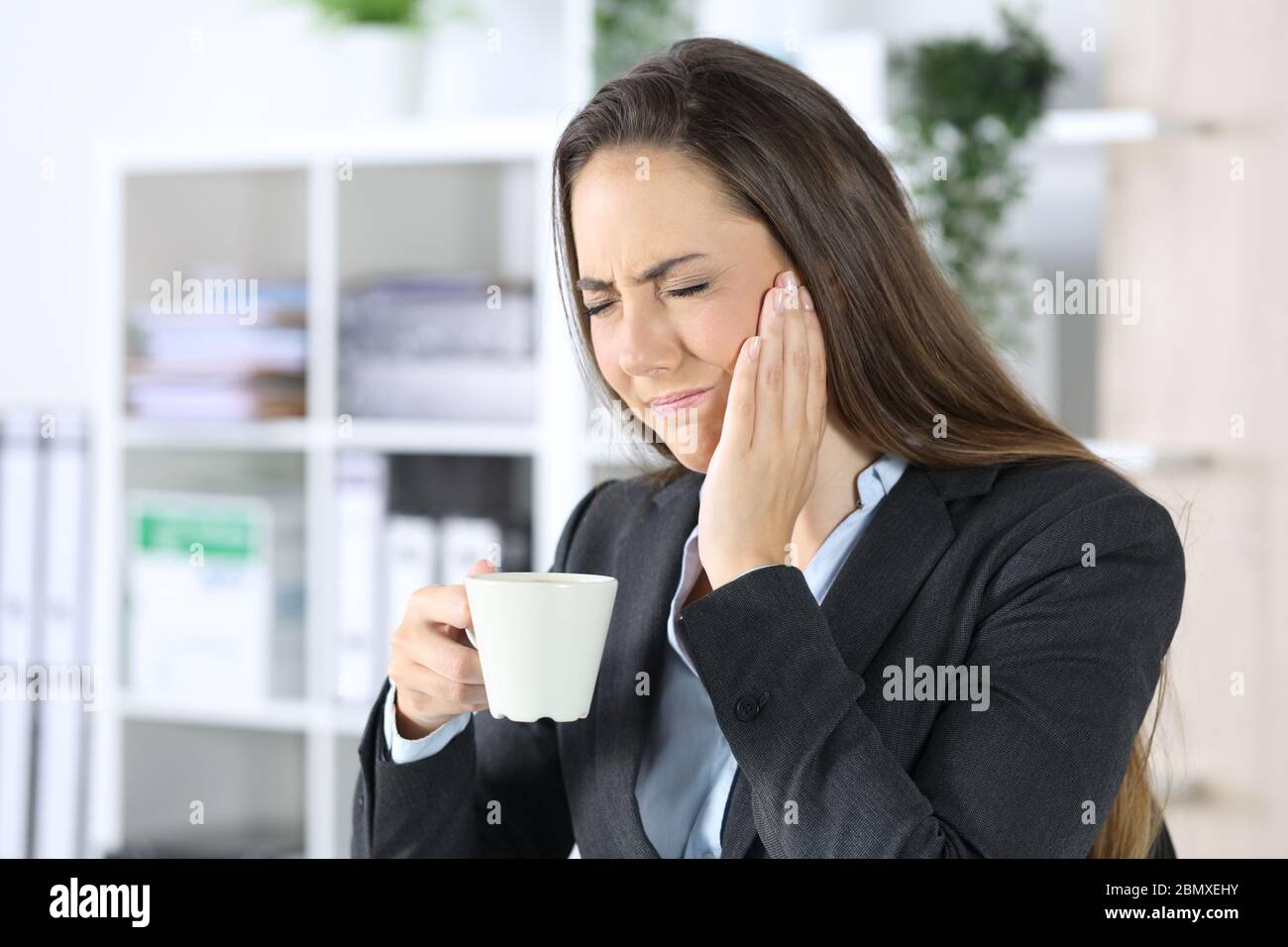 Femme exécutive en douleur avec mal de dents tenant une tasse de café assis au bureau Banque D'Images