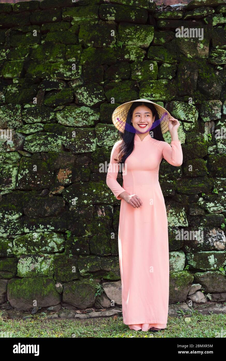 Une femme vietnamienne portant une robe Ao Dai rose et souriant pour la caméra à l'extérieur des murs de mousse d'un temple de la ville de Hue Banque D'Images