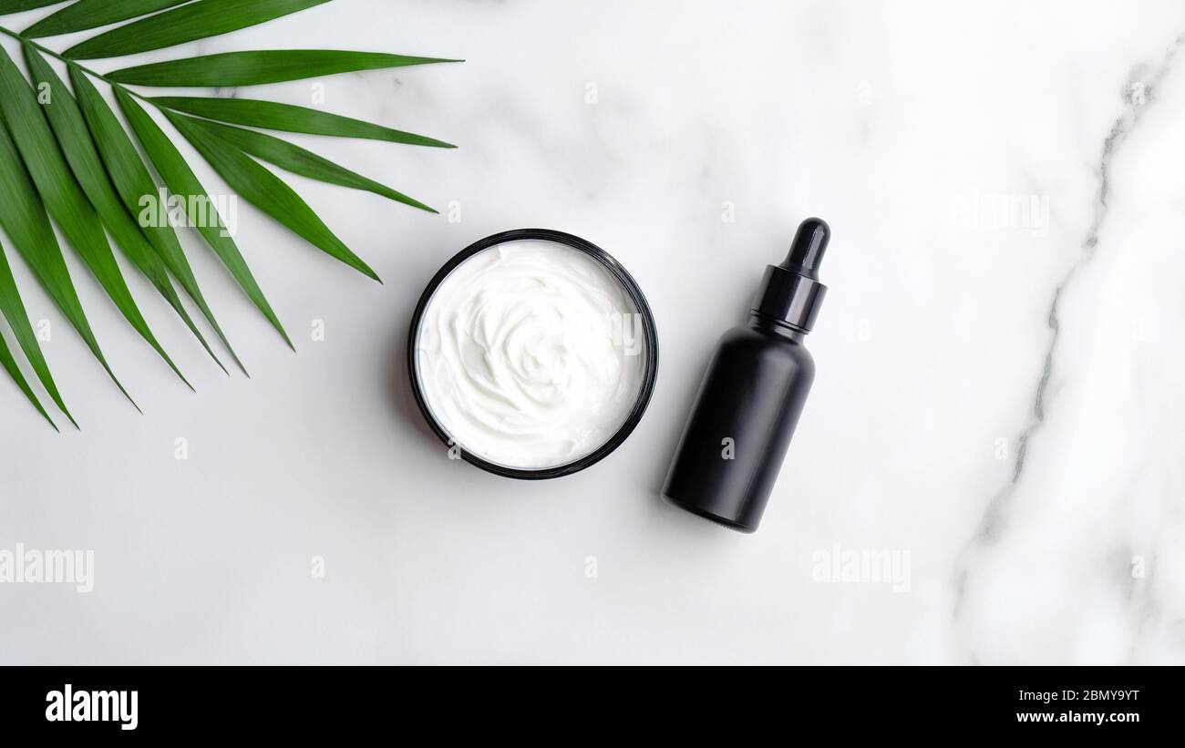 Un gros plan de bouteille d'huile aromatique noire et crème pour la main sur fond de marbre avec des feuilles de palmier. Banque D'Images