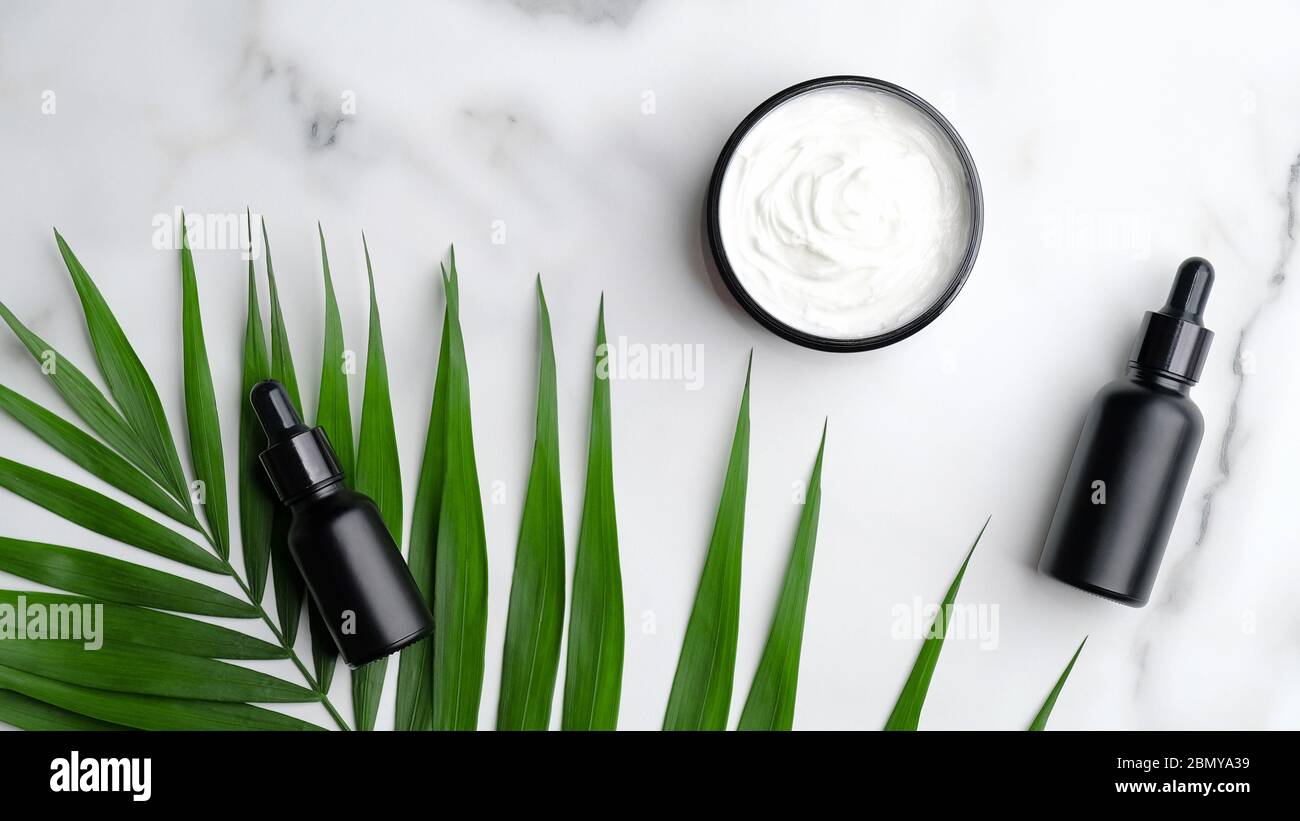 Gros plan de bouteilles d'huile aromatique noire et de crème corporelle sur fond de marbre avec feuilles de palmier. Banque D'Images