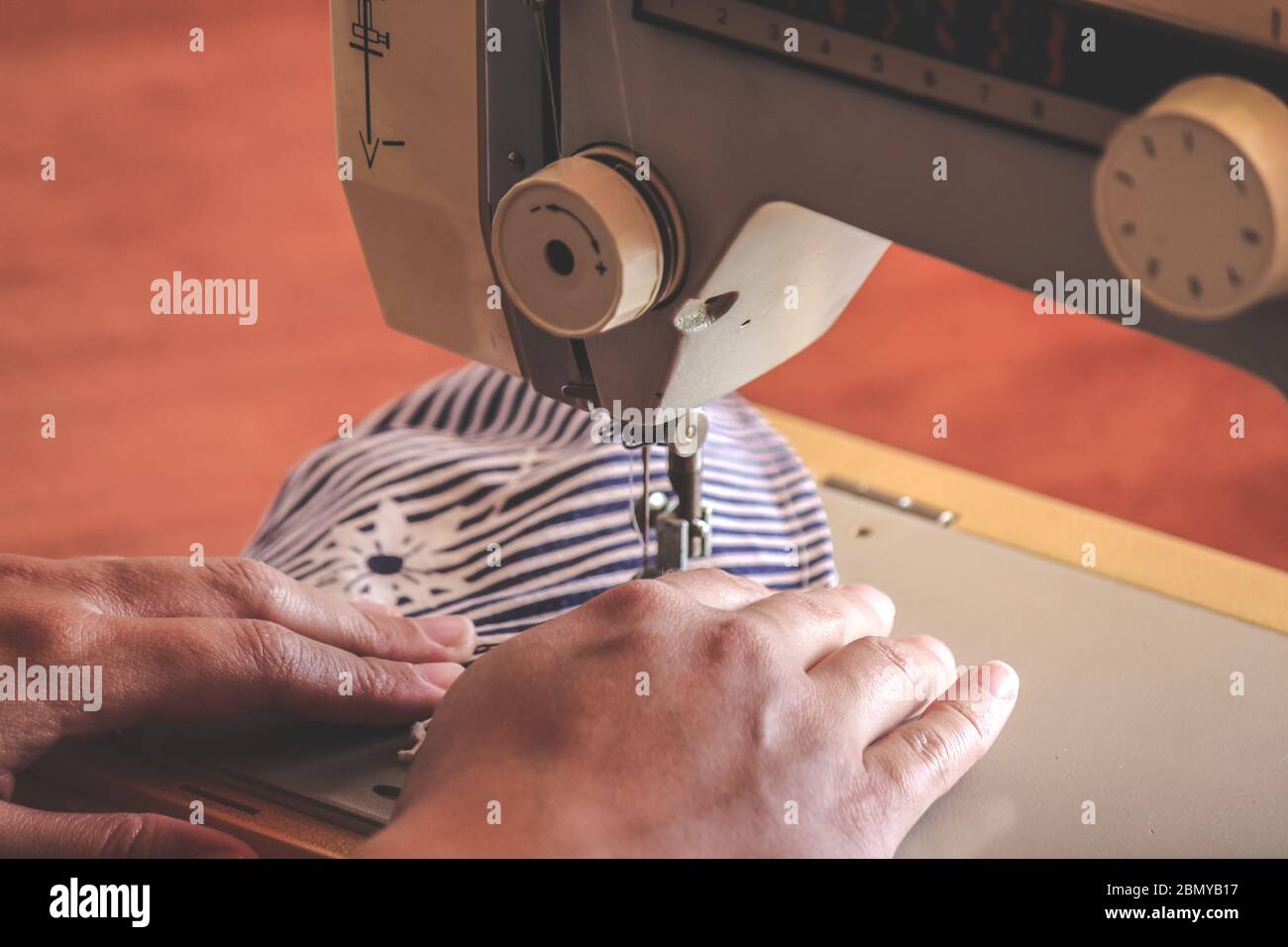 Les mains des femmes utilisant la machine à coudre pour coudre un masque médical coloré du visage pendant la pandémie de coronavirus. Masque de protection fait maison contre le virus. Masques à coudre, COVID-19. Mise au point sélective. Banque D'Images