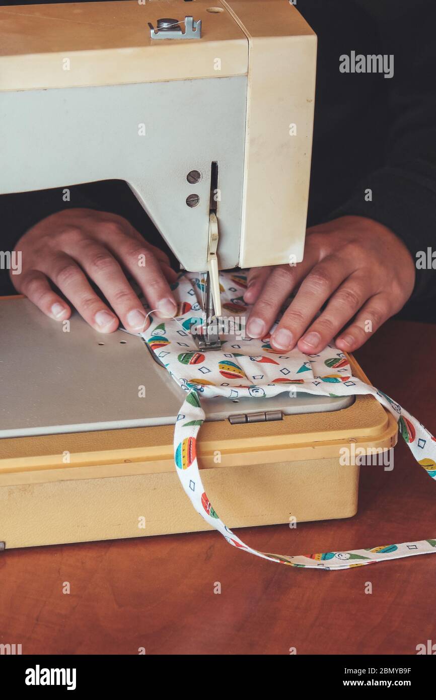 Les mains des femmes utilisant la machine à coudre pour coudre un masque médical coloré du visage pendant la pandémie de coronavirus. Masque de protection fait maison contre le virus. Masques à coudre, COVID-19. Mise au point sélective. Banque D'Images