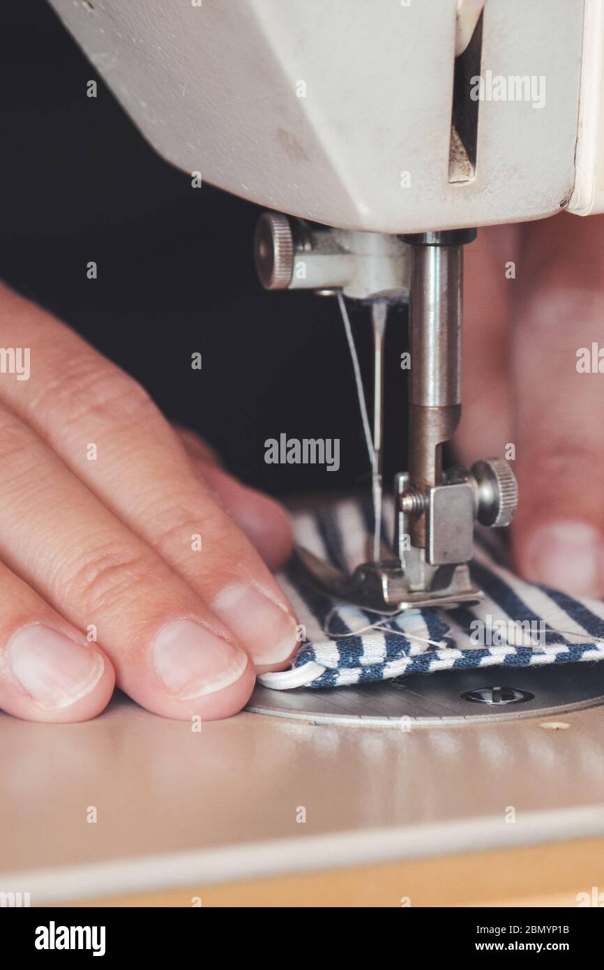Les mains des femmes utilisant la machine à coudre pour coudre un masque médical coloré du visage pendant la pandémie de coronavirus. Masque de protection fait maison contre le virus. Masques à coudre, COVID-19. Mise au point sélective. Banque D'Images