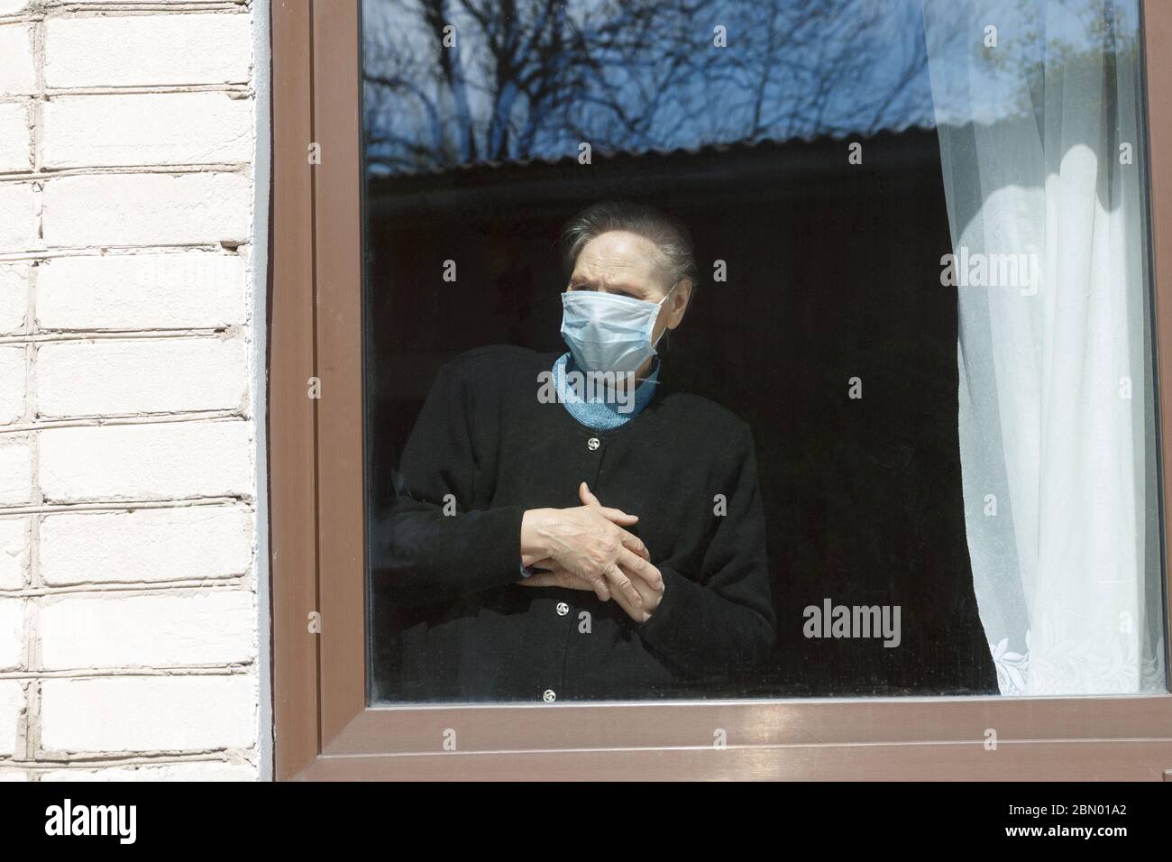 Une vieille grand-mère dans un masque de protection regarde la fenêtre à l'auto-isolation. Banque D'Images