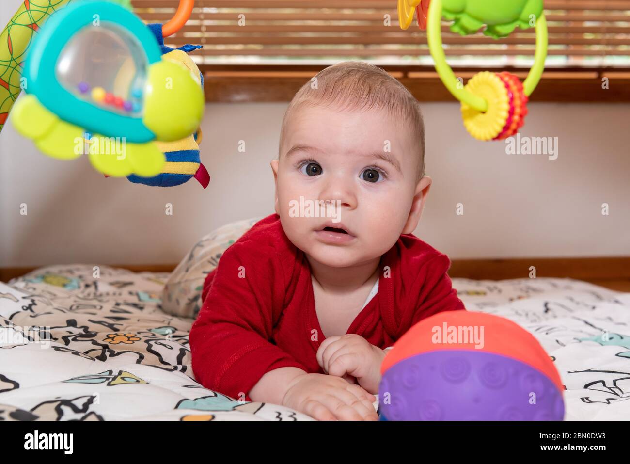 Un petit garçon mignon pendant le ventre, regardant l'appareil photo. enfant de 6 mois avec expression de curiosité sur son visage entouré de jouets colorés. Banque D'Images