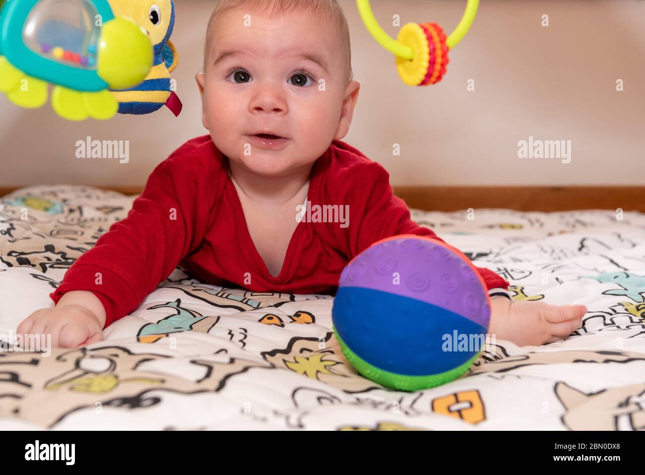 Un petit garçon mignon pendant le ventre, regardant l'appareil photo. enfant de 6 mois avec expression de curiosité sur son visage entouré de jouets colorés. Banque D'Images