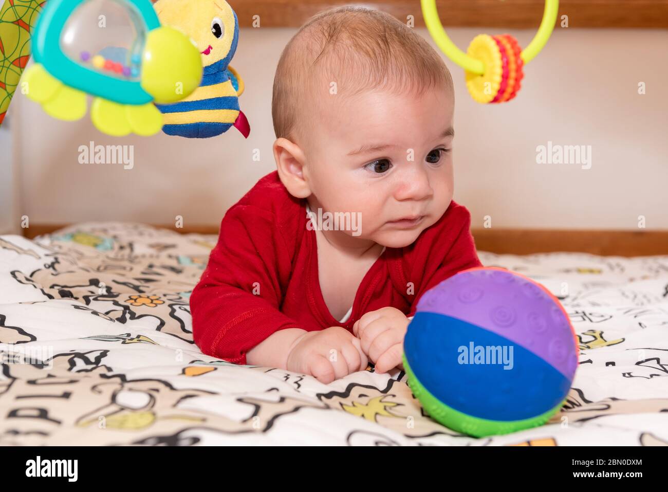 Un petit garçon mignon pendant le ventre, regardant l'appareil photo. enfant de 6 mois avec expression de curiosité sur son visage entouré de jouets colorés. Banque D'Images