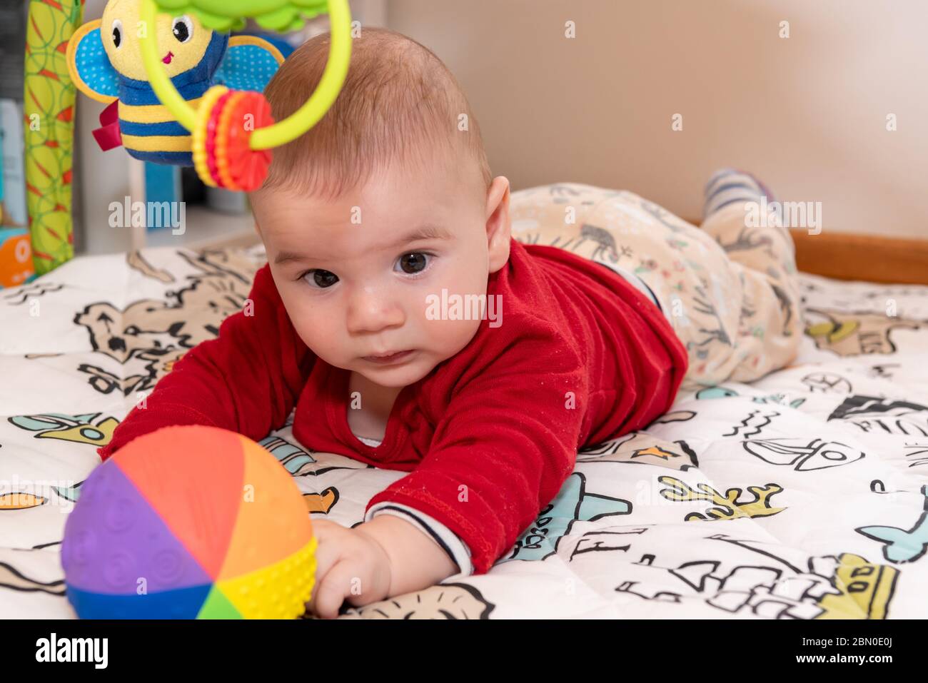 Un petit garçon mignon pendant le ventre, regardant l'appareil photo. enfant de 6 mois avec expression de curiosité sur son visage entouré de jouets colorés. Banque D'Images