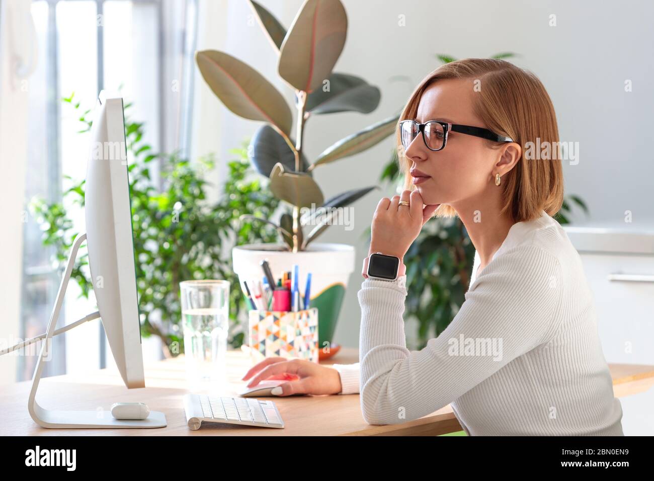 Femme pigiste pensive dans des lunettes travaillant sur l'ordinateur à partir du bureau à domicile pendant la quarantaine en raison du coronavirus. Espace de travail confortable entouré de plantes. Activé Banque D'Images