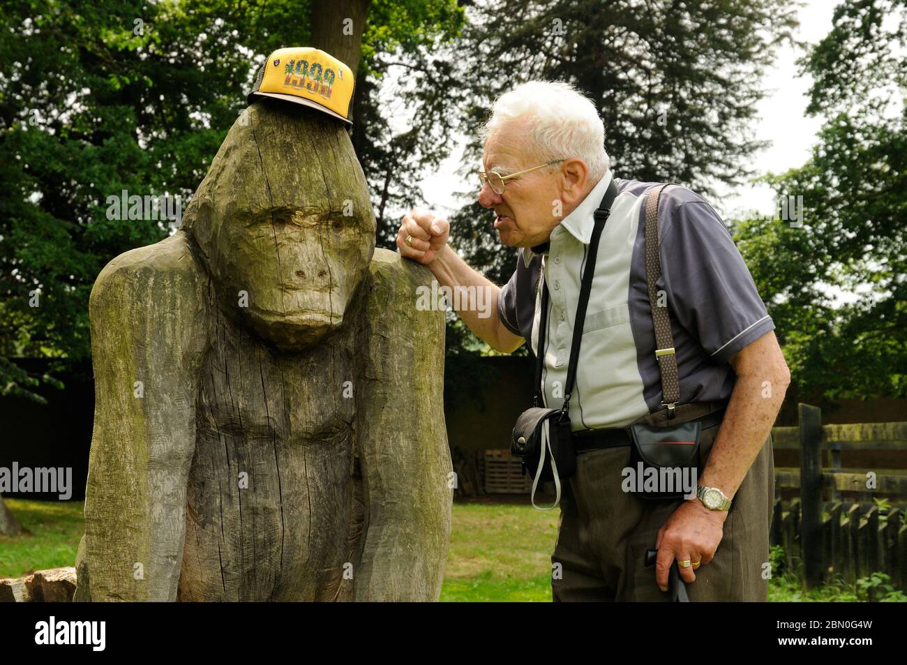 Homme âgé s'amusant avec sa casquette de baseball et un gorille en bois sculpté au parc de campagne d'Elsham Hall, Lincolnshire, Angleterre Banque D'Images