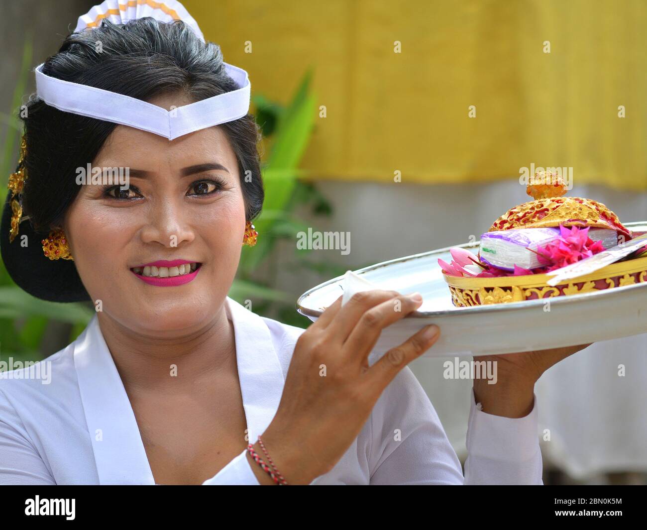 La femme indonésienne balinaise en blanc offre des offrandes lors d'une cérémonie religieuse de temple hindou et sourit pour la caméra. Banque D'Images