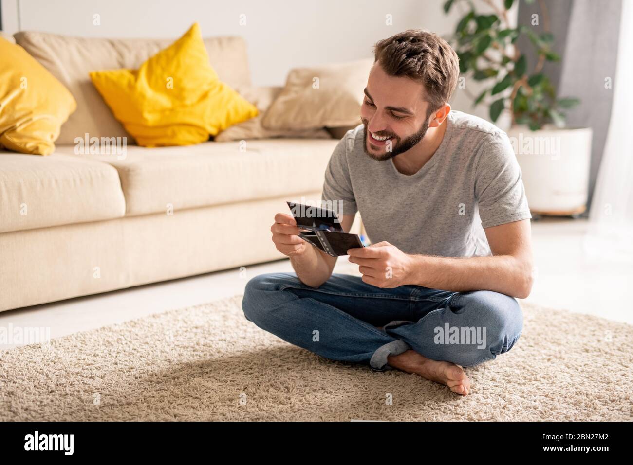 Jeune homme joyeux et barbu assis avec des jambes croisées sur la moquette et en appréciant la nostalgie tout en regardant de vieilles photos dans le salon pendant l'auto-isolation Banque D'Images