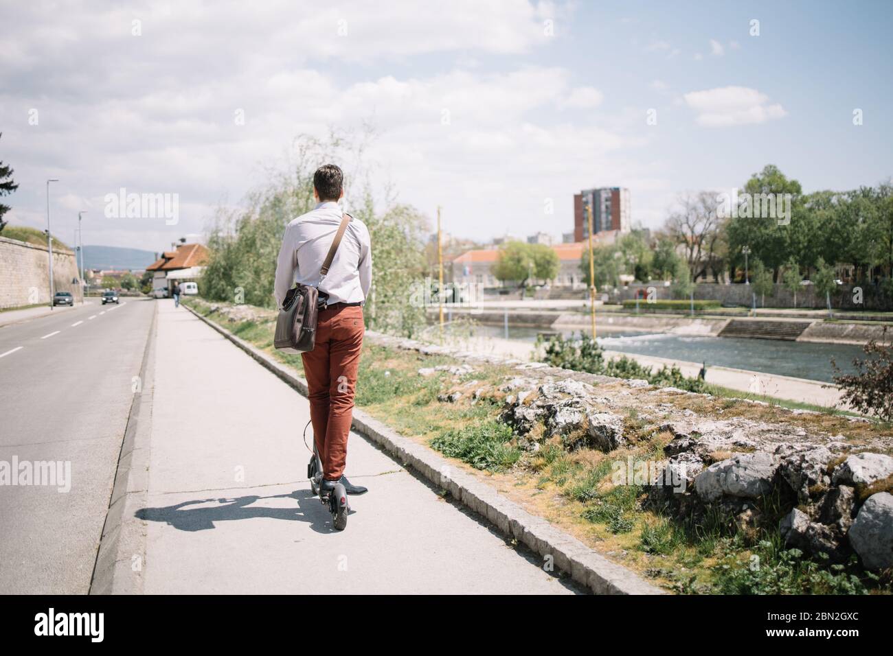 Dos de l'homme sur un scooter électrique conduisant sur la route au bord de la rivière. Vue arrière de l'homme d'affaires portant une chemise et un sac en conduisant le scooter sur le trottoir. Banque D'Images