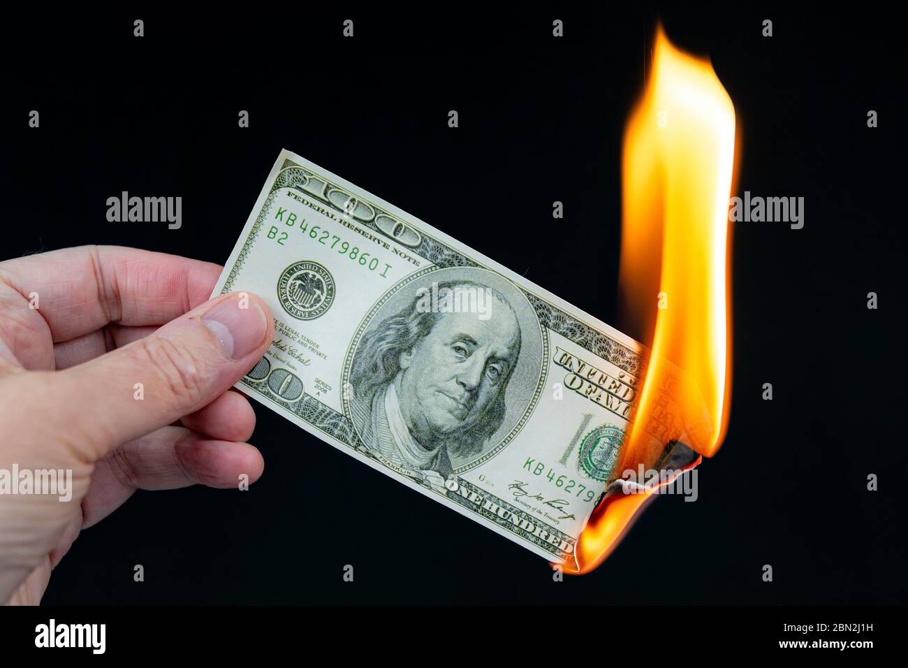 Brûler une facture de 100 $ US. Une main tient le billet de banque en feu. Environ un quart de celui-ci est consommé par une grande flamme. Arrière-plan noir. Banque D'Images