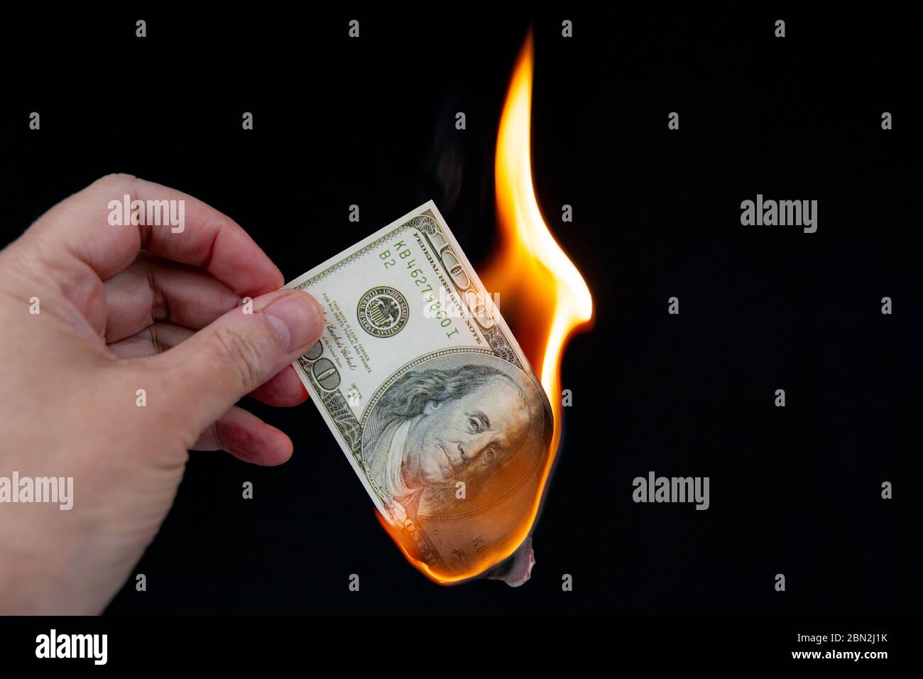 Brûler une facture de 100 $ US. Une main tient le billet de banque en feu, environ la moitié a été brûlée. Arrière-plan noir. Banque D'Images