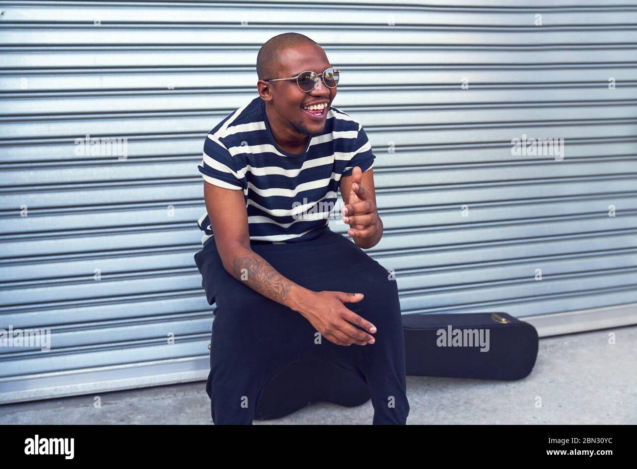 Joyeux musicien masculin assis sur une caisse de guitare à l'extérieur du garage Banque D'Images