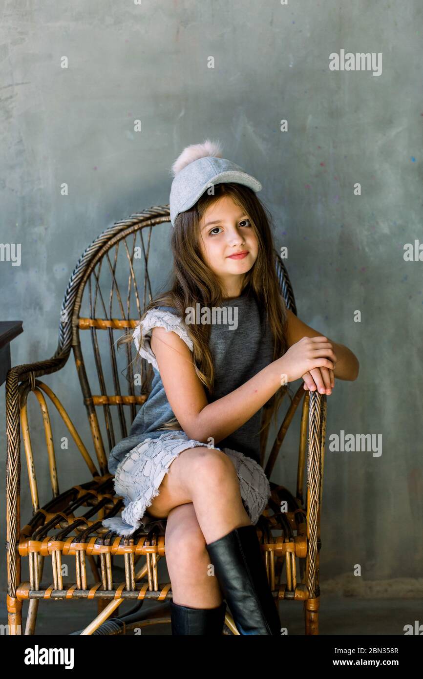 Belle petite fille aux cheveux longs bruns et aux yeux bleus assis dans un fauteuil habillé de robe grise et un chapeau Banque D'Images