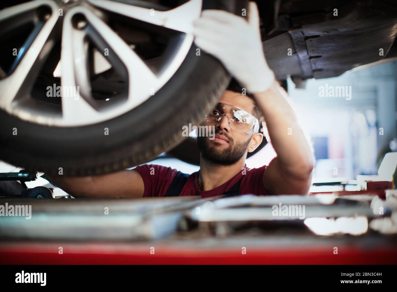 Mécanicien de sexe masculin travaillant sous voiture, examinant les pneus dans un atelier de réparation automobile Banque D'Images
