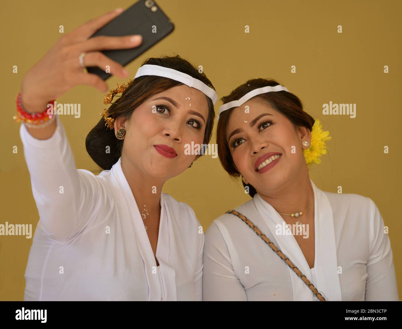Deux belles femmes balinaises indonésiennes habillées en blanc prennent une photo de la wefie avec leur téléphone cellulaire lors d'une cérémonie religieuse hindoue au temple. Banque D'Images